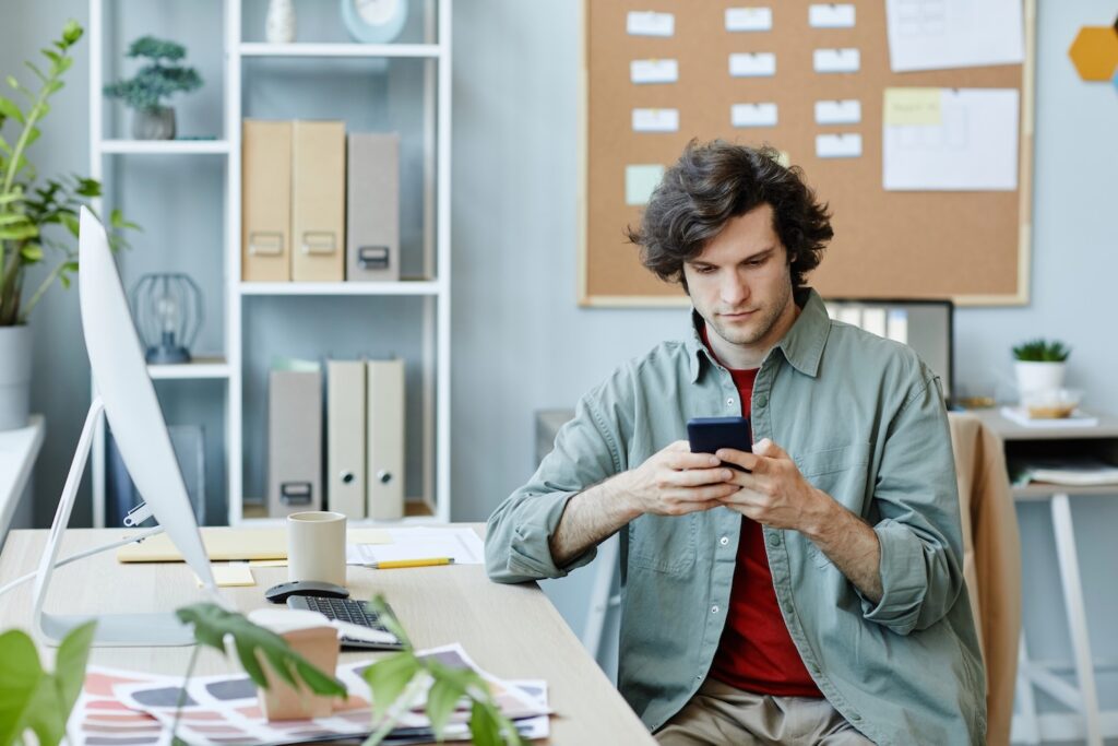 Portrait of Caucasian young man using smartphone at workplace and scrolling social media during break