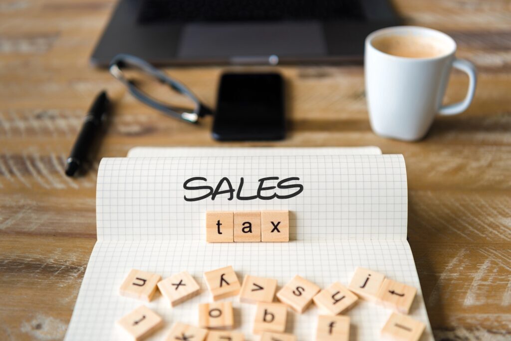 Closeup on notebook over vintage desk surface, front focus on wooden blocks with letters making Sales Tax text. Business concept image with office tools and coffee cup in background