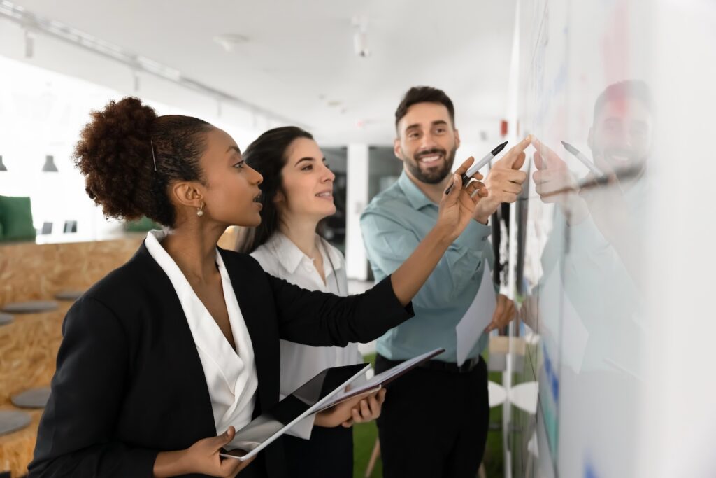 Diverse teammates brainstorm ideas, writing them on white board, collaboratively planning project or strategy, discussing, pointing to elements on board, review visual representation of plan or data