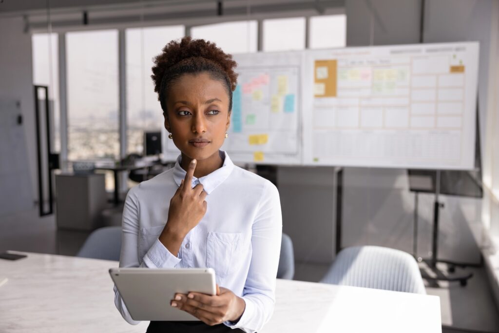 Serious young African American businesswoman holding tablet, thinking on online project electronic report, standing with digital gadget in office alone, looking away, touching chin, making decision